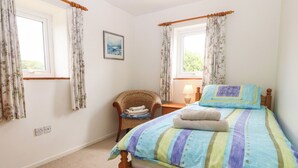 A bedroom with a bed, two windows with curtains, a chair, a bedside table with a lamp, and a wall-mounted picture. - Porth Awyr (Pwllheli)