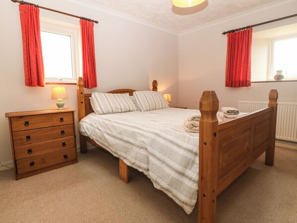 A bedroom with a wooden bed, a wooden dresser with drawers, two bedside lamps, and red curtains. - Porth Awyr (Pwllheli)