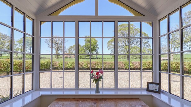 Une véranda dotée de grandes fenêtres, d’un banc en bois et d’un vase rempli de fleurs.
