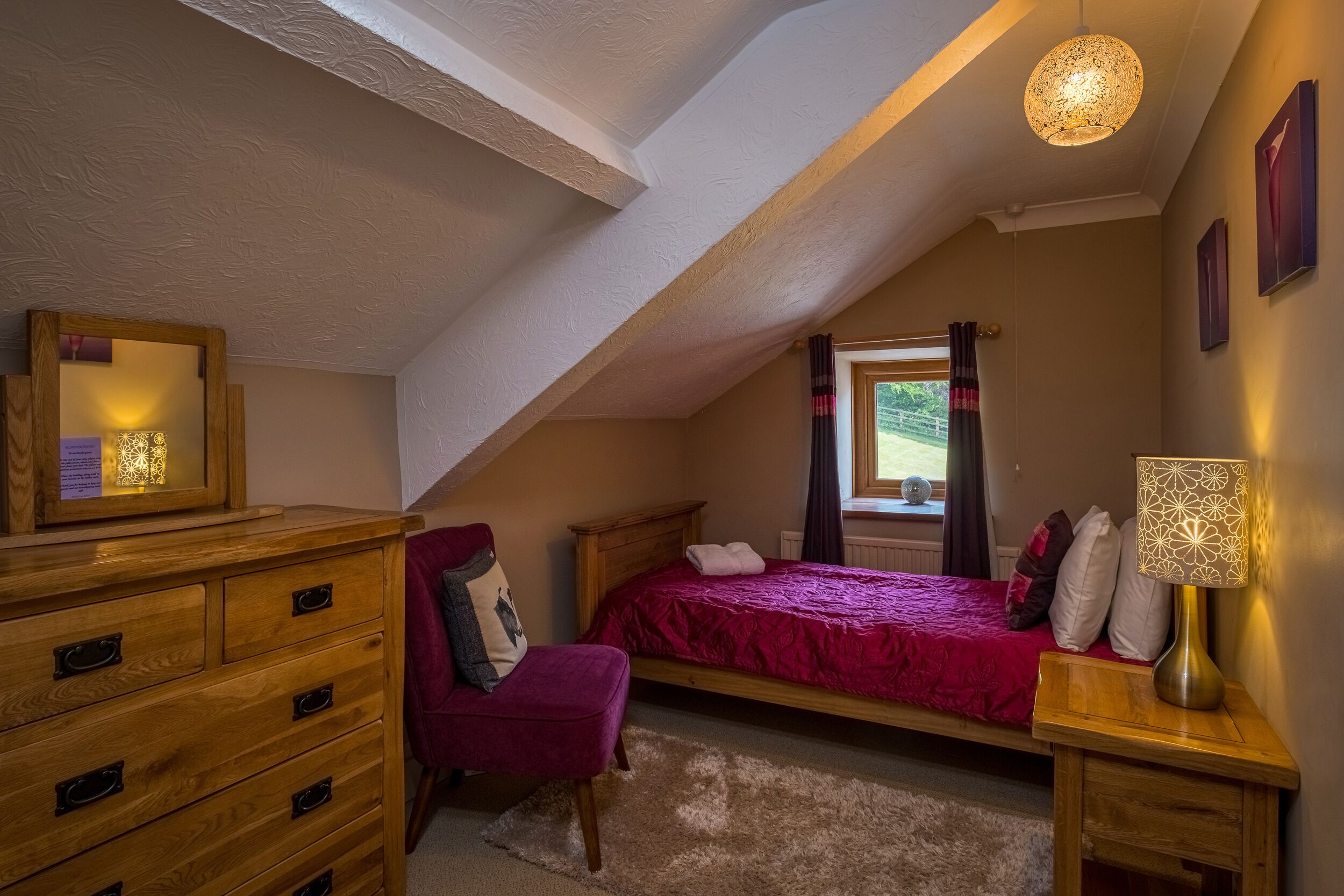 A bedroom with a sloped ceiling, a wooden dresser, a red bed, a chair, a nightstand, and a lamp.