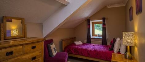 A bedroom with a sloped ceiling, a wooden dresser, a red bed, a chair, a nightstand, and a lamp.