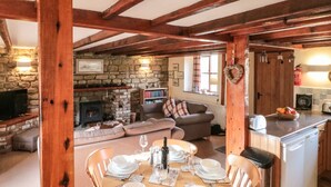 Uma sala de jantar com mesa de madeira posta para quatro pessoas, lareira de pedra, sala de estar com sofá e estantes, e cozinha com torradeira.