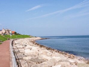 A coastal walkway with a stone barrier, a clear sky, and a distant view of a building with a red roof. - Roma (Colwyn Bay)
