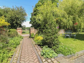 A brick pathway leading to a garden area with a metal gate, surrounded by lush greenery and a stone wall. - Just A Cottage, Newlands Farm (Alfreton)