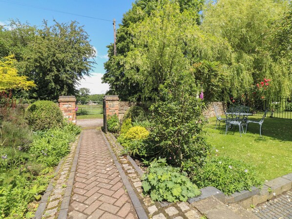 A brick pathway leading to a garden area with a metal gate, surrounded by lush greenery and a stone wall. - Just A Cottage, Newlands Farm (Alfreton)