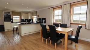 A modern kitchen with a central island, a dining area with a wooden table and black chairs, and large windows with curtains. - Little Laight (Stranraer)