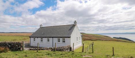 A white house with a grey roof, surrounded by a stone wall and a fence, set in a rural landscape with a body of water in the background.