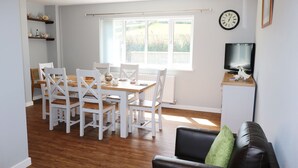 A dining area with a wooden table and white chairs, a black leather sofa, a television on a wooden stand, and a window with a view of greenery. - Pen Y Cae (Denbigh)