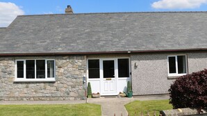 A stone house with a grey roof, a white door, and a small garden with a bush and a potted plant. - Pen Y Cae (Denbigh)