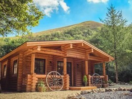 Una cabaña de troncos con un porche, dos ruedas grandes y montañas al fondo.
