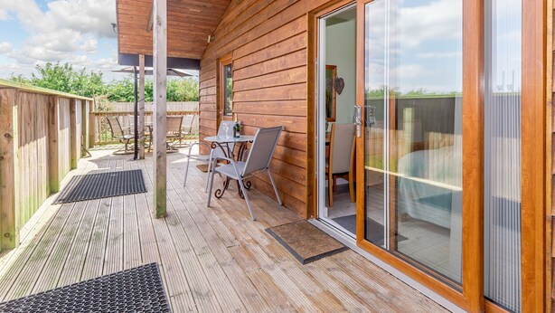 A wooden deck with a table and chairs, a bottle of champagne, and a sliding glass door leading to an outdoor area. - Kingfisher Lodge (Driffield)