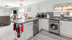 A modern kitchen with a dishwasher, washing machine, and a built-in oven. There is a dining area with a table and chairs, and a living room with a sofa. - Kingfisher Lodge (Driffield)