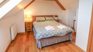 Une chambre avec un plafond en pente, une tête de lit en bois, un lit avec une literie à motifs floraux, une table de chevet avec une lampe et un radiateur.