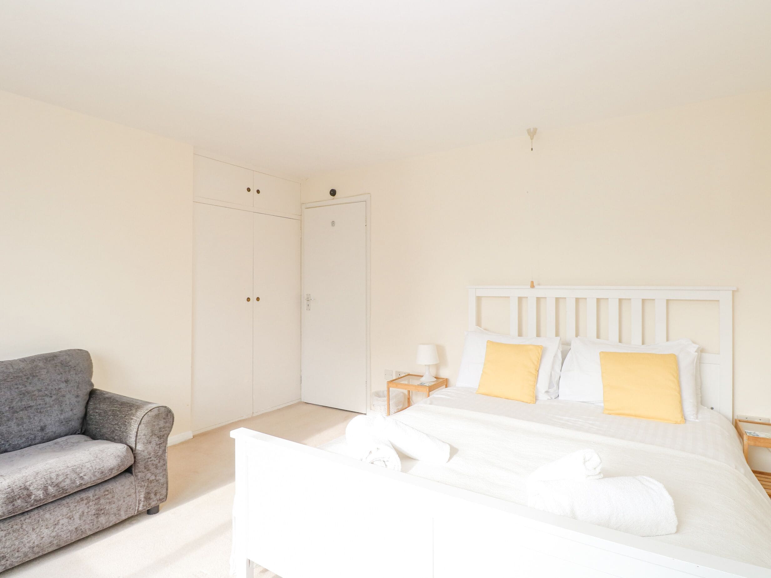 A bedroom with a bed, a grey sofa, a wardrobe, and a nightstand with a lamp.