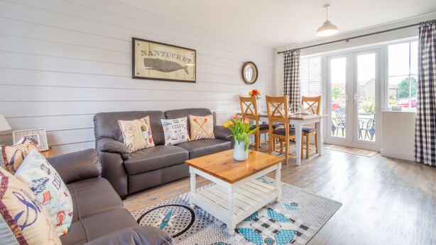 A living room with a brown leather sofa, a wooden coffee table, and a dining area with a table and chairs. There is a large window with checkered curtains, a framed picture on the wall, and a vase with flowers on the coffee table. - Lilipad Cottage (Filey)