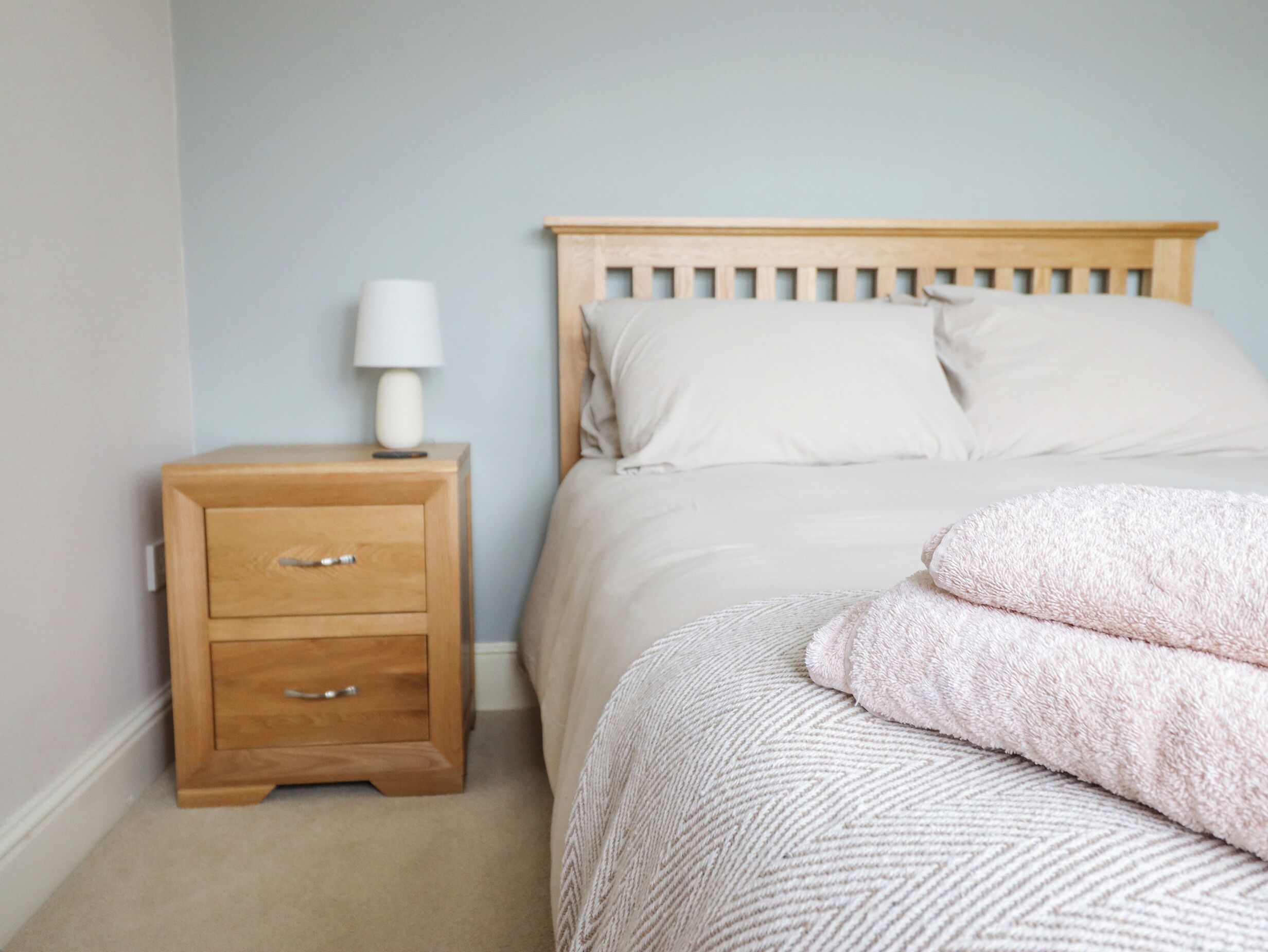 Un dormitorio con una mesita de noche de madera, una lámpara, una cama con ropa de cama blanca y una toalla rosa.
