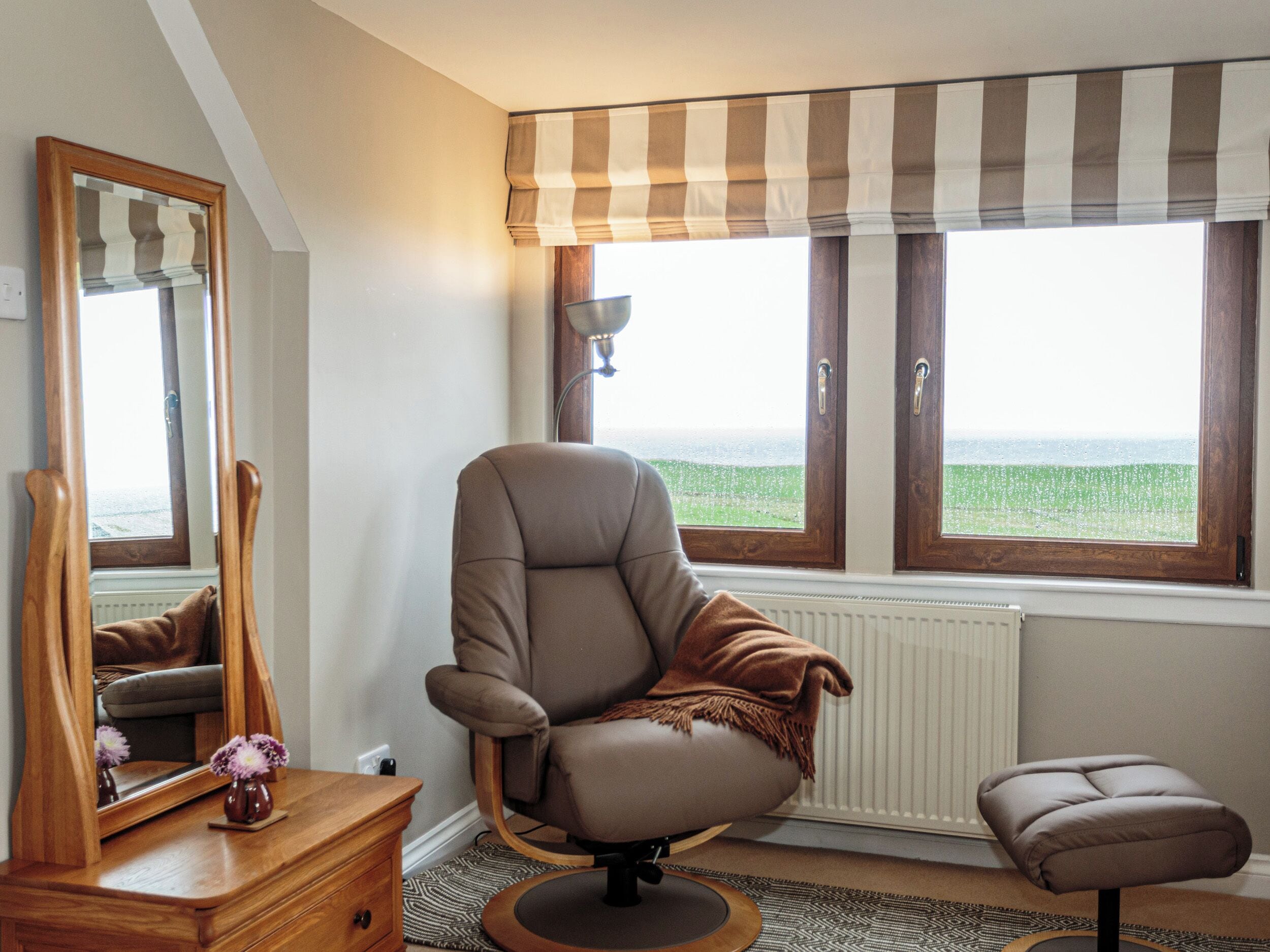 A room with a large window, a chair, a small stool, a mirror, and a wooden dresser.