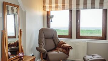 A room with a large window, a chair, a small stool, a mirror, and a wooden dresser.