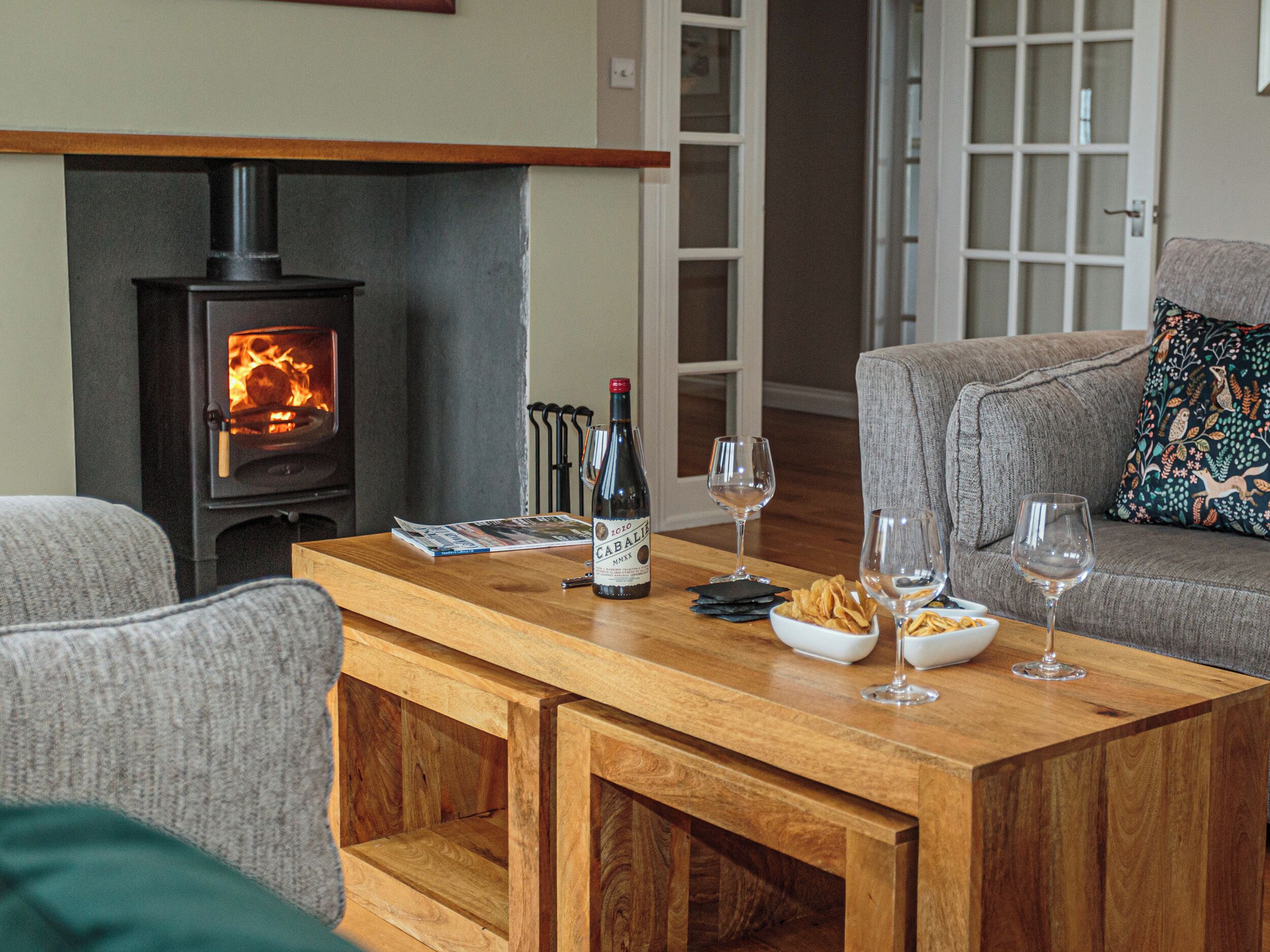 Ein gemütliches Wohnzimmer mit Kamin, einem Holztisch mit Zeitschriften, Weingläsern und Snacks.