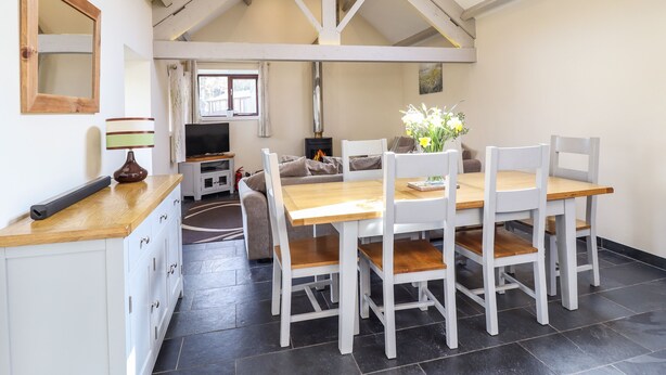 A dining area with a wooden table and white chairs, a TV on a stand, and a fireplace in the background. - Beudy Bach Barn (Bala)