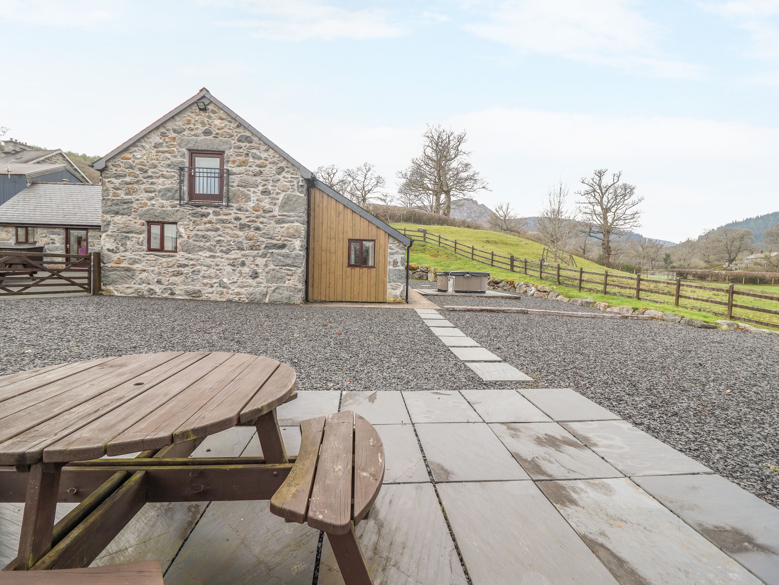 A stone building with a wooden door and windows, a gravel patio with a wooden table, and a fenced area with a view of hills.