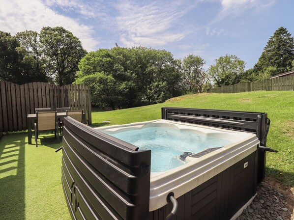 A hot tub with a wooden deck and chairs, surrounded by a grassy yard and a wooden fence.