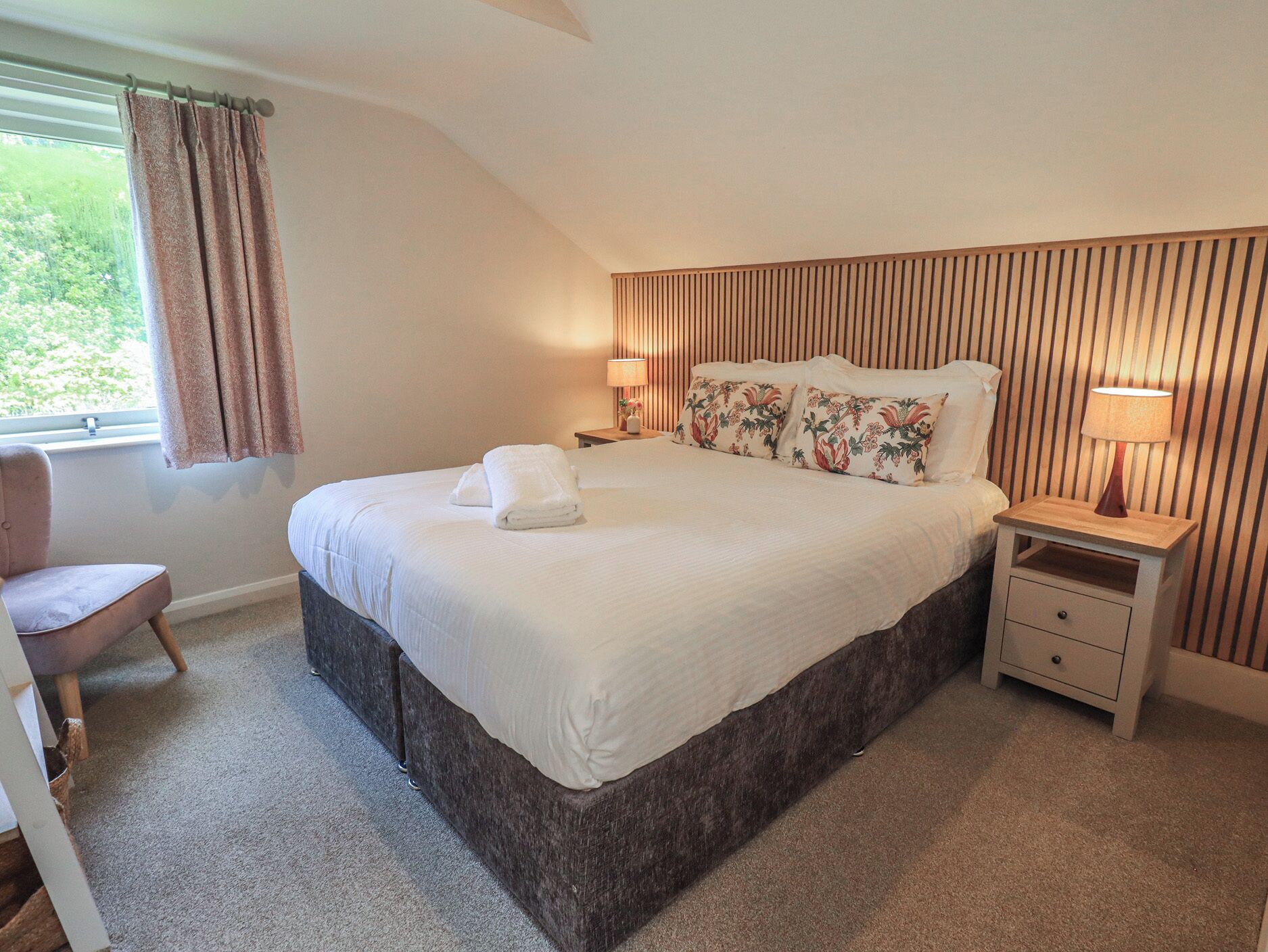 A bedroom with a large bed, two bedside tables, a chair, a window with curtains, and a sloped ceiling.
