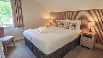 A bedroom with a large bed, two bedside tables, a chair, a window with curtains, and a sloped ceiling.