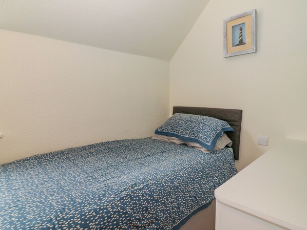 A small, neatly made bed with a blue and white patterned bedspread and matching pillow, a wooden headboard, and a framed picture of a lighthouse on the wall. - Nathaniel's Cottage (Kirkcudbright)