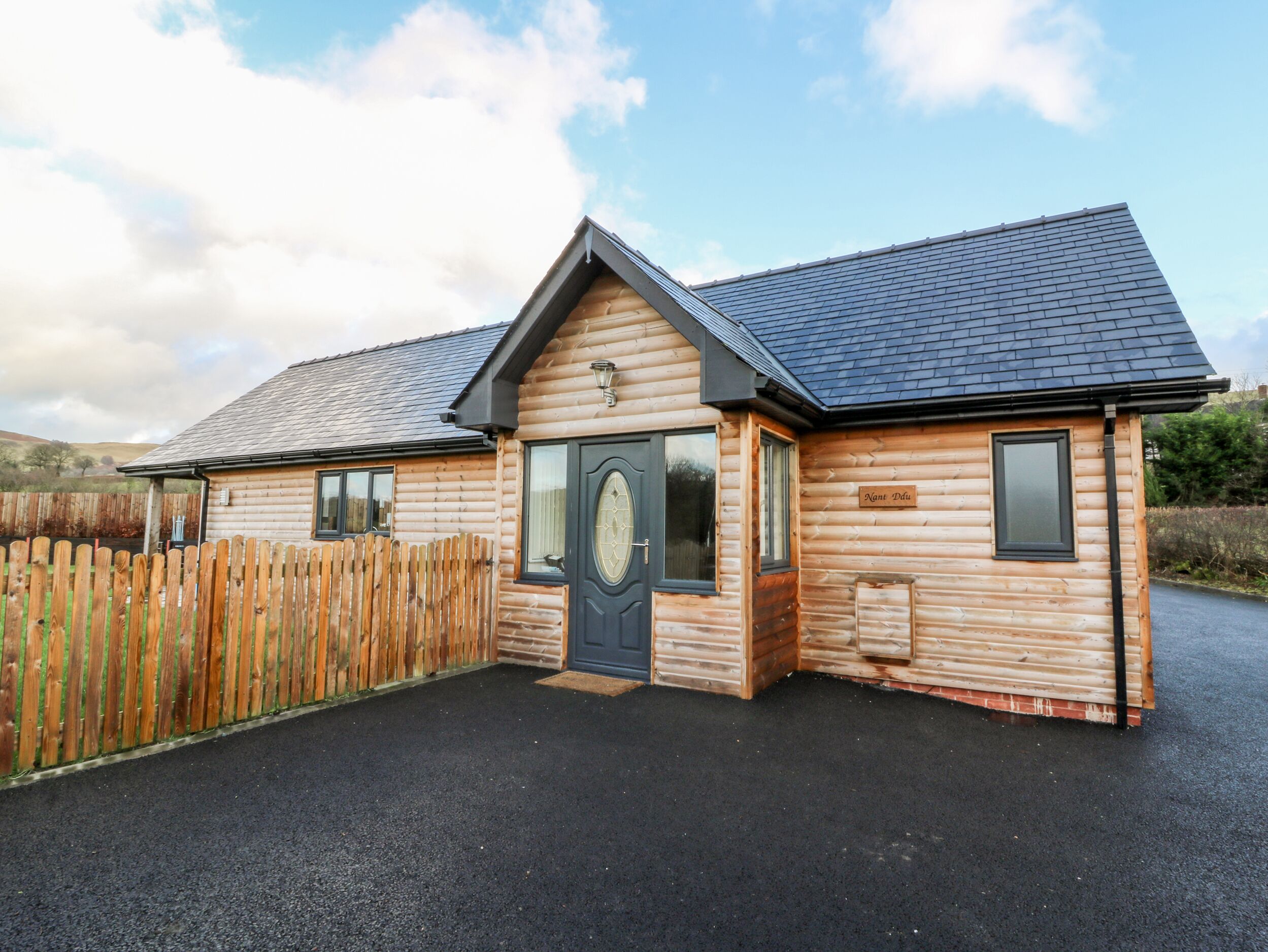 A log cabin with a dark roof, a front door with a glass panel, and a sign reading "Nant Pudu".