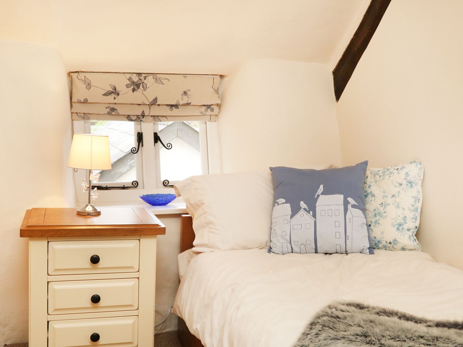 Un dormitorio con una cama, una mesita de noche con lámpara, una ventana con cortina floral, y un estante de pared con un cuenco azul.