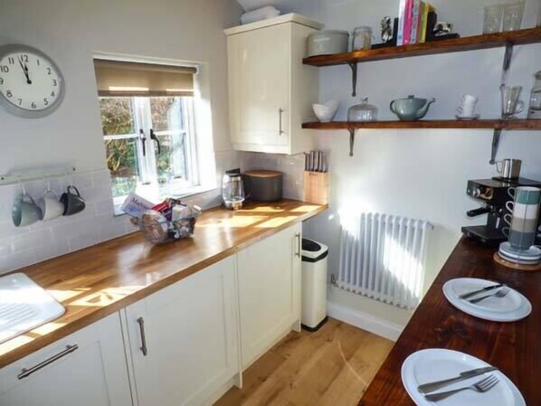 A kitchen with white cabinets, a wooden countertop, a coffee machine, and a window with blinds. - Kitts Cottage (Malvern)