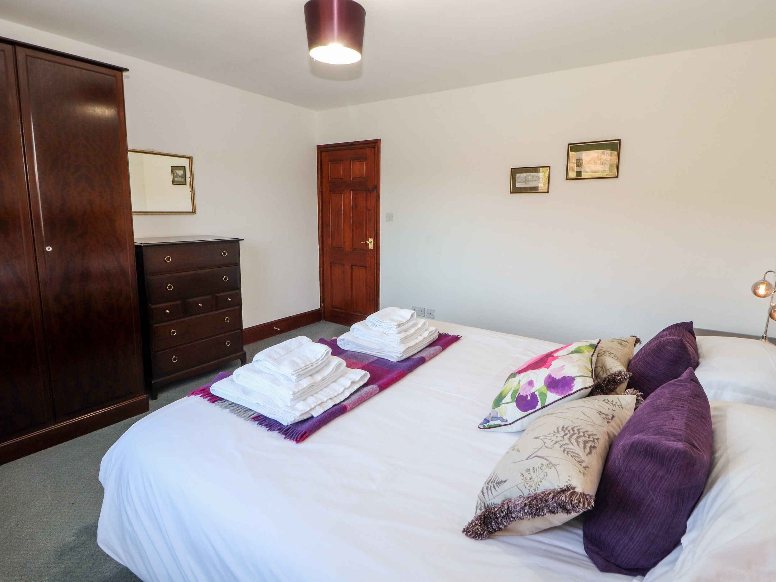 A neatly made bed with white linens and a purple throw blanket, a wooden wardrobe, a dresser, and two framed pictures on the wall.