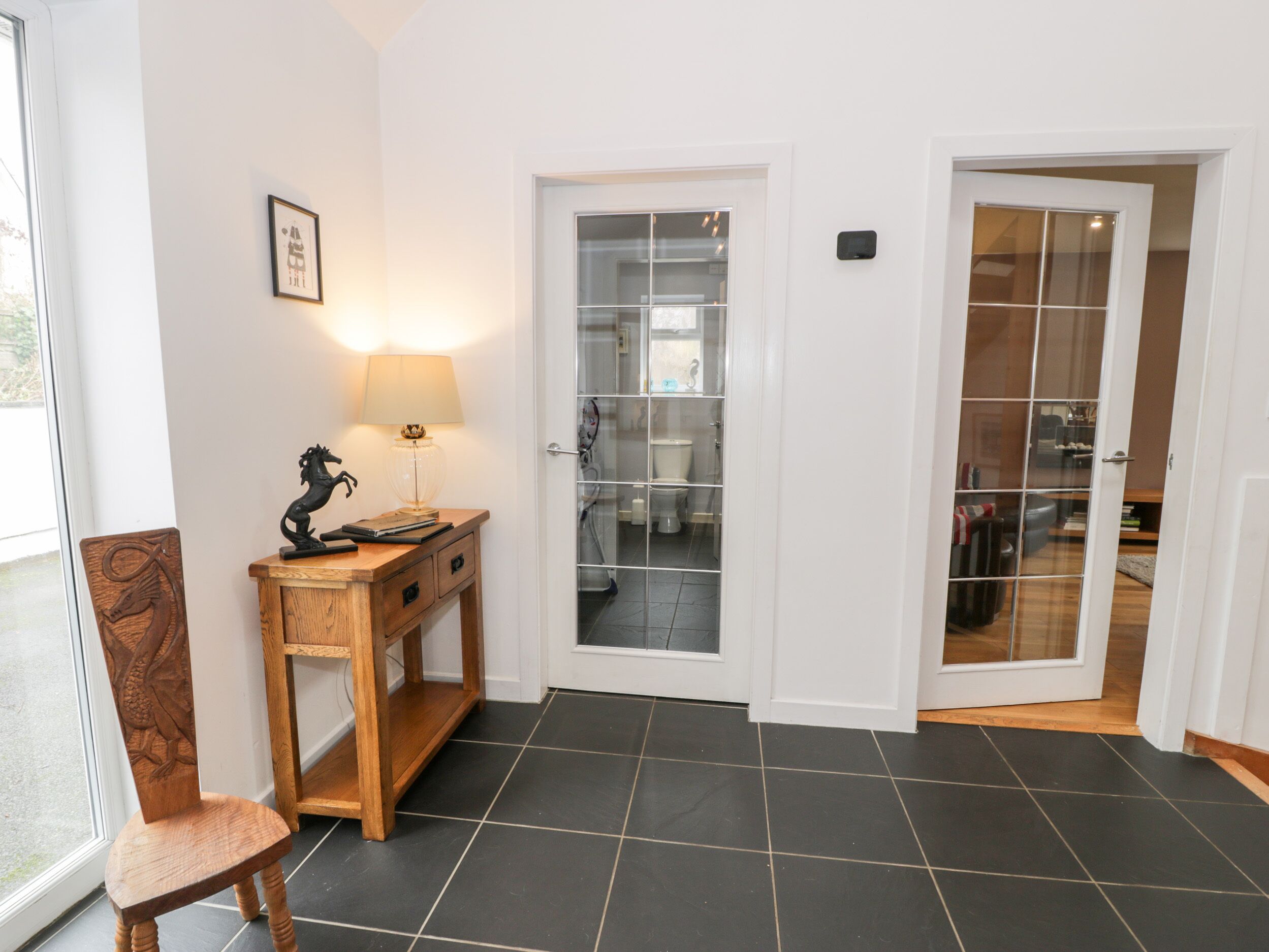 A hallway with a wooden desk, a chair, and a lamp. There is a door leading to a bathroom with a glass partition.