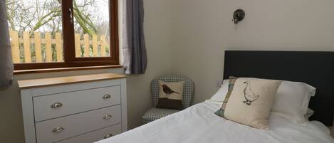 A bedroom with a bed, a nightstand, a dresser, a window with curtains, and a wall-mounted light.