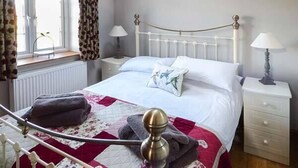 Ein Schlafzimmer mit weißem Metallbettgestell, einer gesteppten Bettdecke, zwei Nachttischen mit Lampen, einem Fenster mit Vorhängen und einem Heizkörper.