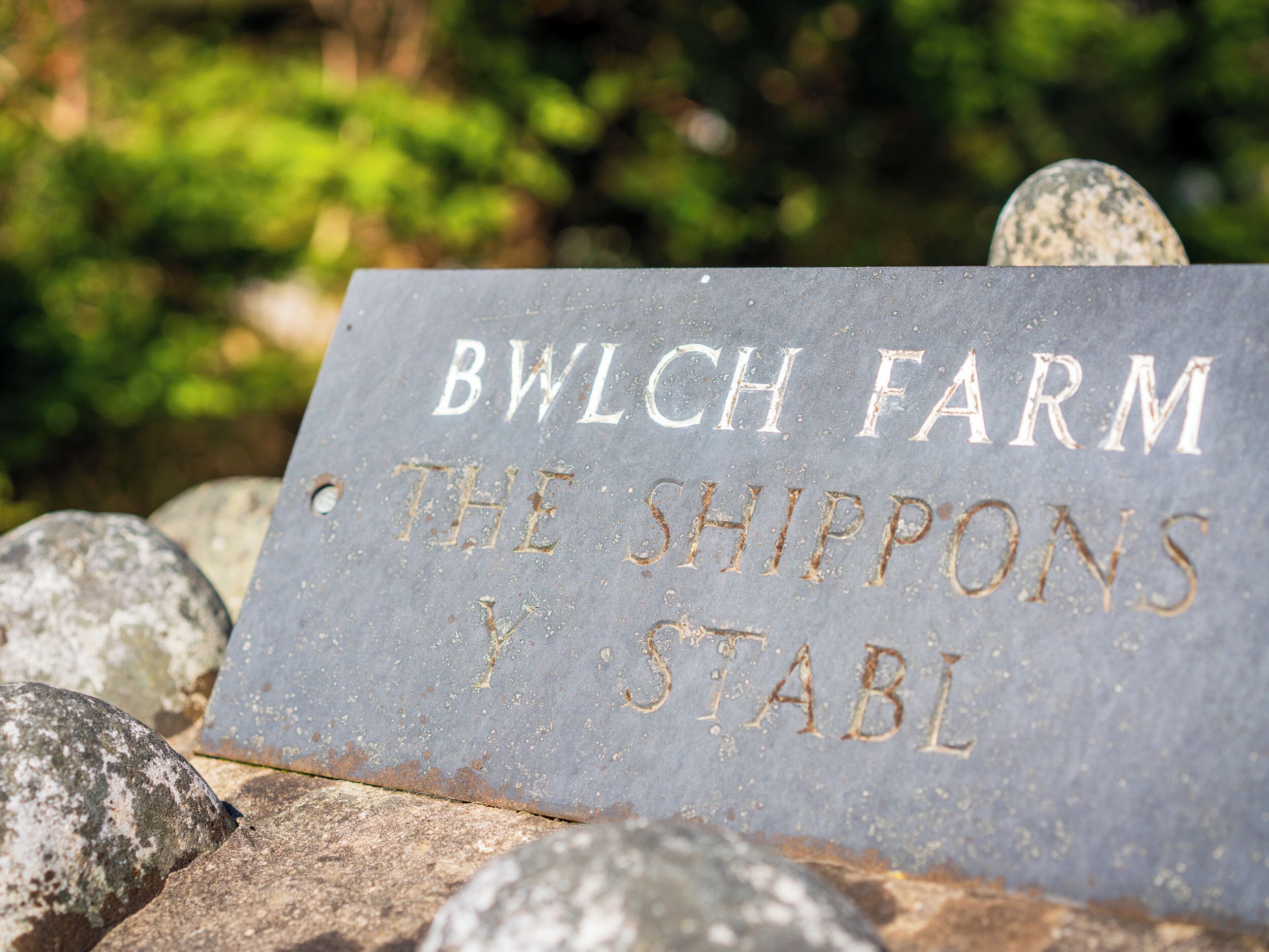 A sign with the text "BWLCH FARM THE SHIPPONS Y STABL" on a stone surface.