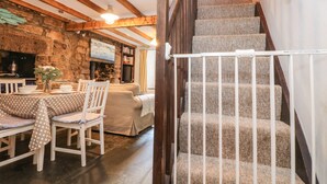 A cozy interior with a stone fireplace, wooden beams, a dining area with a table and chairs, and a staircase with a white railing.