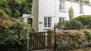A white stone cottage with a wooden gate, surrounded by greenery.