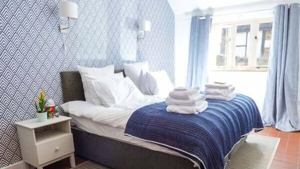 Una cama bien tendida con sábanas blancas y una manta azul, una mesita de noche con una lámpara, y una pared con estampado.
