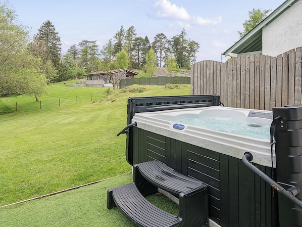 A hot tub with a wooden deck and steps, surrounded by a wooden fence and a grassy yard. - Maple - Woodland Cottages (Windermere)
