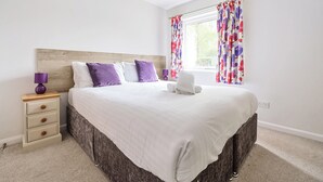 A neatly made bed with white bedding and purple pillows, a wooden headboard, a bedside table with a lamp, and a window with floral curtains. - Maple - Woodland Cottages (Windermere)