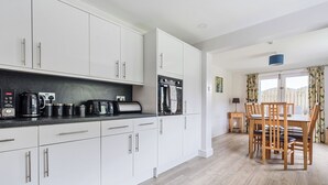 A modern kitchen with white cabinets, a coffee maker, toaster, and microwave. There is a dining area with a wooden table and chairs. - Maple - Woodland Cottages (Windermere)