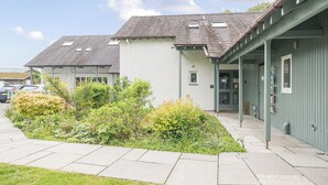A building with a sloped roof, a covered porch, and a small garden area with various plants. - Maple - Woodland Cottages (Windermere)