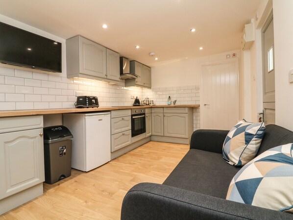 A compact kitchen with a built-in oven, a toaster, and a coffee maker. There is a grey sofa with patterned cushions in the living area. - 2 Moorlands (Minehead)