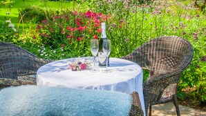 Una mesa redonda con mantel blanco, dos copas de vino y una botella de vino rosado, rodeada de sillas de mimbre en un jardín.