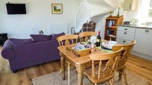 A living room with a purple sofa, wooden dining table with chairs, a kitchen area with cabinets, and a staircase. - Gwdihw (Oswestry)