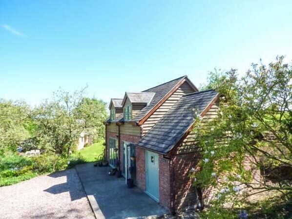 A small, two-story wooden house with a sloped roof, a front porch, and a gravel driveway. - Gwdihw (Oswestry)