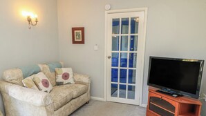 Una sala de estar con un sofá color beige, un televisor de pantalla plana sobre un mueble de madera, una puerta blanca con paneles de vidrio azul y una lámpara fijada a la pared.