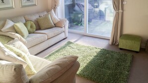 A living room with a beige sofa, green and white cushions, a green rug, and a sliding glass door leading to a backyard.